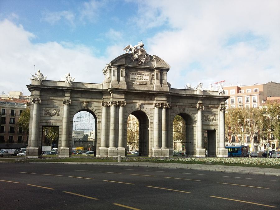 La Puerta de Alcalá se alza como uno de los grandes símbolos de Madrid, testigo del tránsito constante de la ciudad y de su propia historia. Construida en el siglo XVIII por orden de Carlos III, su arquitectura monumental permanece casi intacta mientras todo cambia a su alrededor. La imagen recoge ese contraste entre permanencia y movimiento, entre la memoria de piedra y la vida contemporánea que continúa atravesándola.
