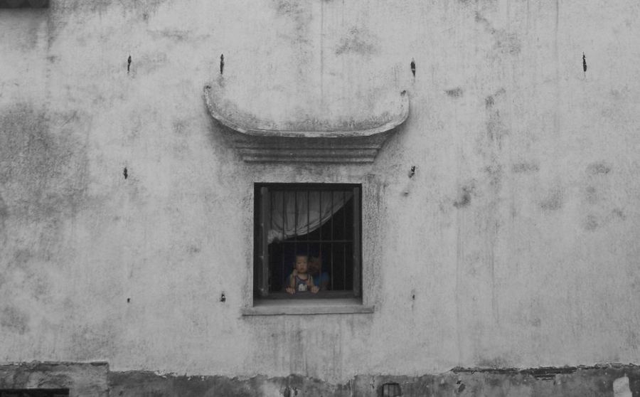 En una antigua calle de Suzhou, una pequeña figura observa el mundo desde detrás de una ventana enrejada. La inmensidad del muro envejecido convierte su presencia en algo casi frágil, mientras la arquitectura tradicional permanece como testigo silencioso. La imagen habla de distancia, curiosidad y de esa frontera invisible entre quien mira y el mundo que continúa fuera.