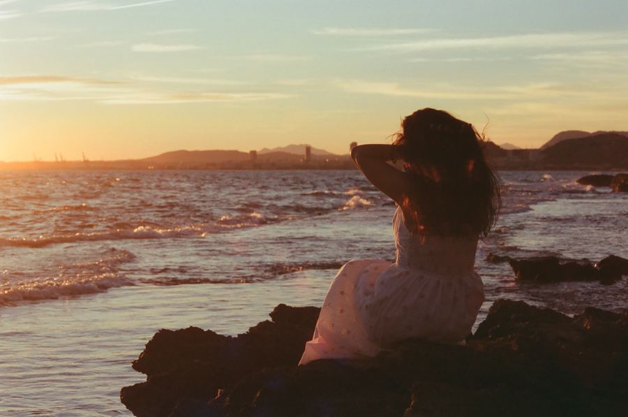 Mujer sentada de espaldas sobre las rocas mirando al mar durante la puesta de sol