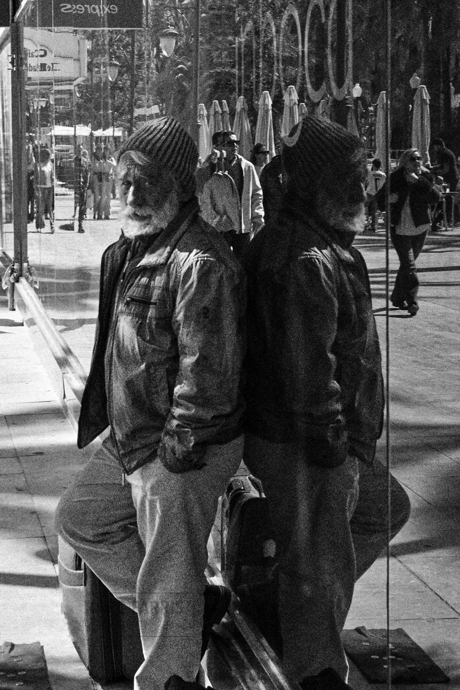 Hombre mayor con gorro apoyado en un escaparate junto a su propio reflejo, donde se refleja el pasado y el futuro del hombre que fue y será. En blanco y negro