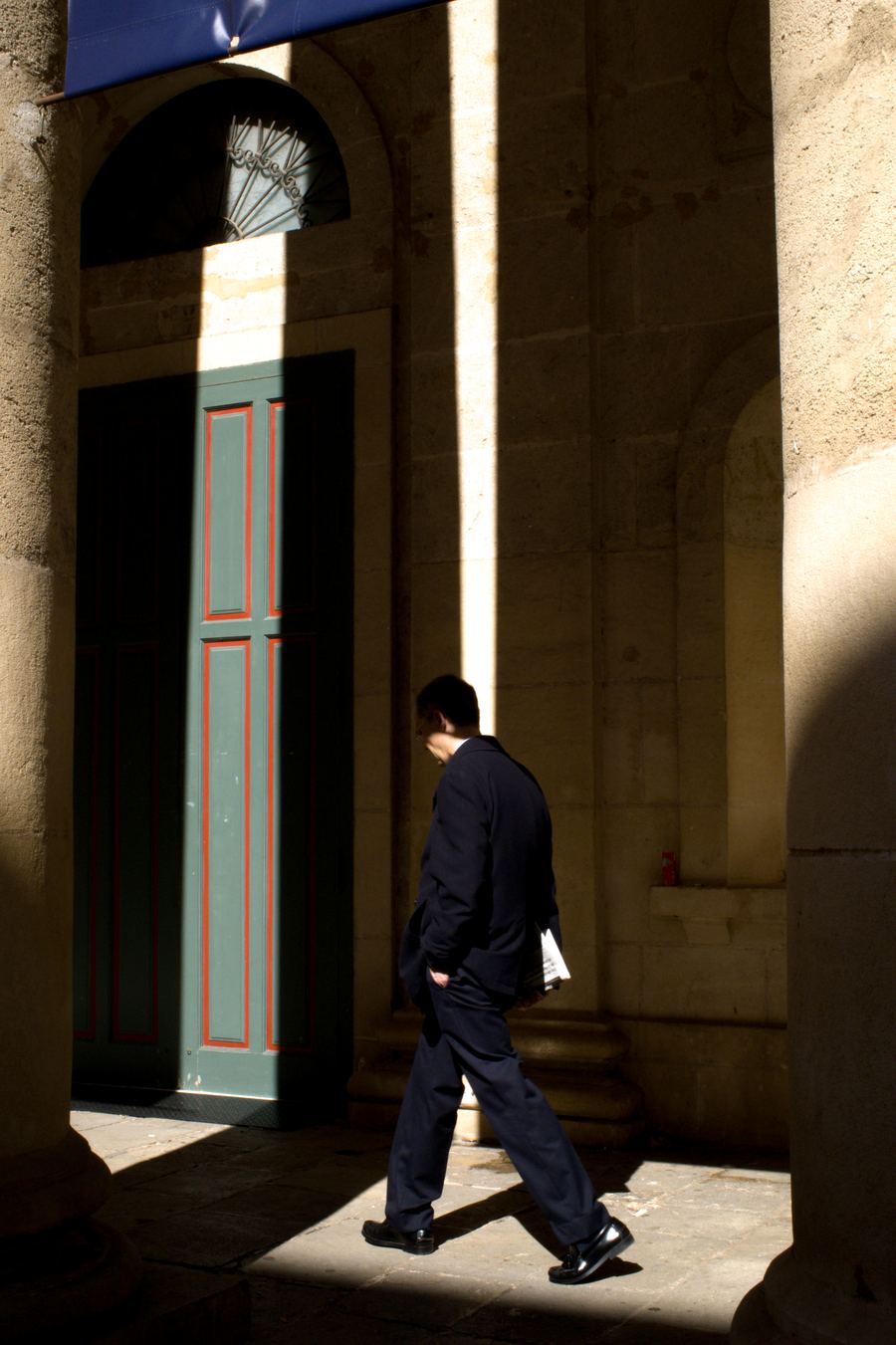 Hombre de traje cruzando un haz de luz junto a una puerta verde y roja bajo unos soportales